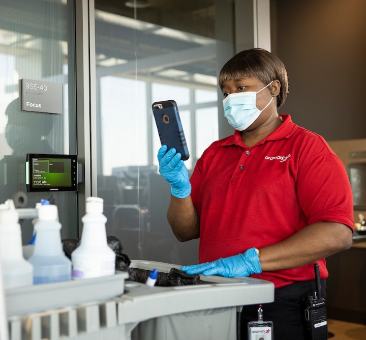 An Aramark worker wearing PPE holding her phone up in front of a cleaning cart in an office.